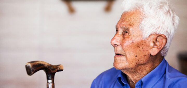 An older man sits and looks up at someone he's talking to. he has white hair, mottled skin, and is wearing a blue shirt. His cane stands nearby.