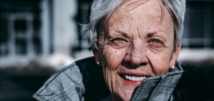 An older woman with short gray hair smiles toward the camera. She appears vital and alive as she peers around the standup collar of her fall coat.